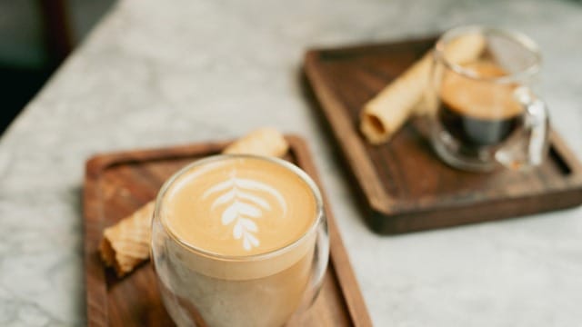 Two coffee cups with latte art and espresso on wooden trays with wafer rolls on a marble table in a cozy cafe setting.