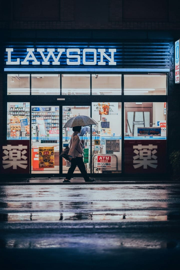 Person with umbrella walking past a brightly lit Lawson convenience store at night, reflection visible on wet pavement.