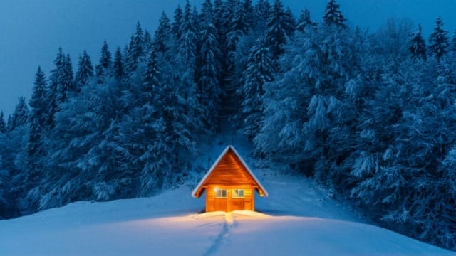 Cozy cabin with warm light in snowy forest, tracks leading to door, surrounded by tall, dark trees in early evening.