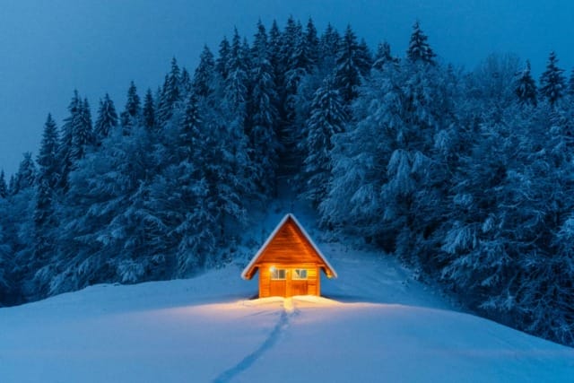 Cozy cabin with warm light in snowy forest, tracks leading to door, surrounded by tall, dark trees in early evening.