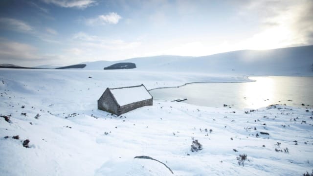 Snowy landscape with a small stone cabin near a frozen lake and mountains under a cloudy sky.