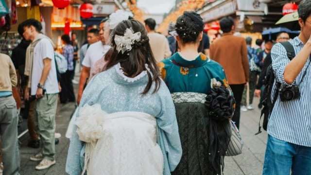 Two people in traditional clothing walking through a crowded street market with colorful decorations and lanterns.