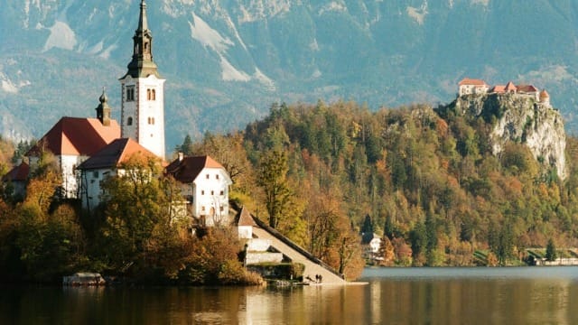 Scenic view of a church by a lake with mountains in the background, reflecting in the calm water.