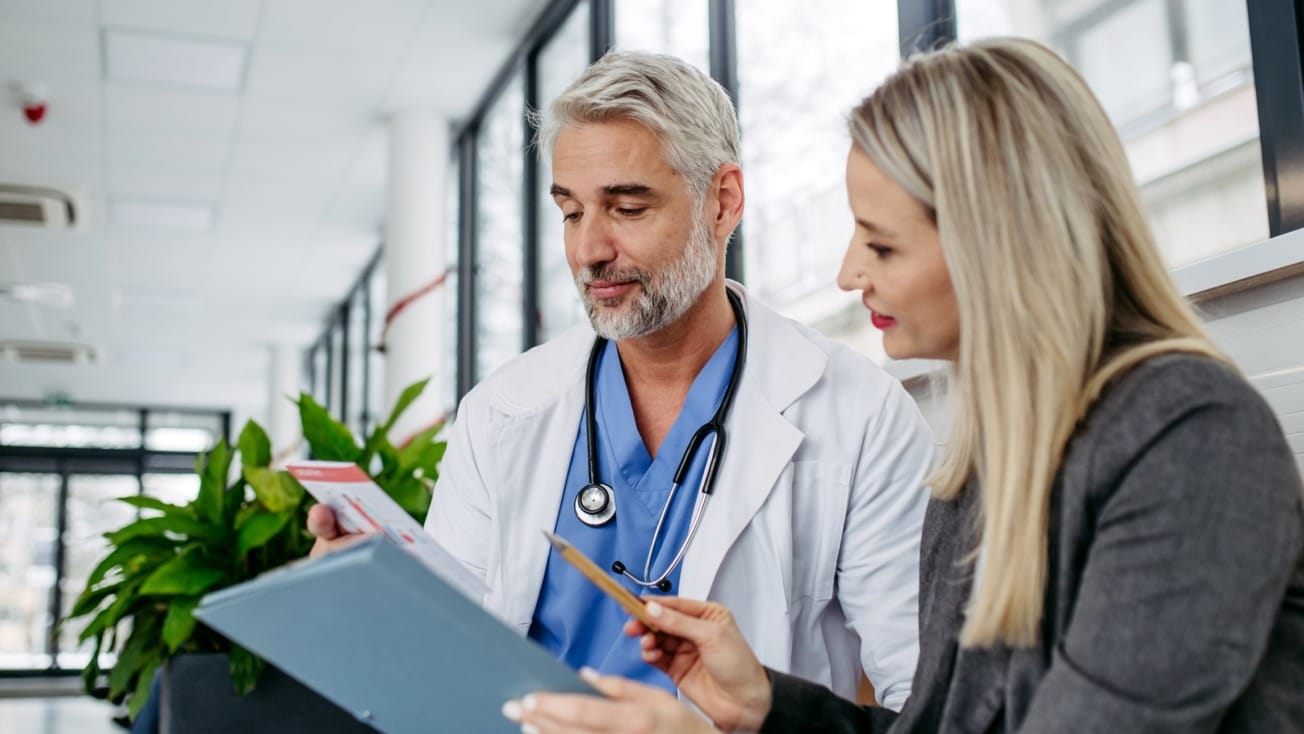 Doctor and woman discussing a medical chart in a bright office setting with large windows and plants in the background.