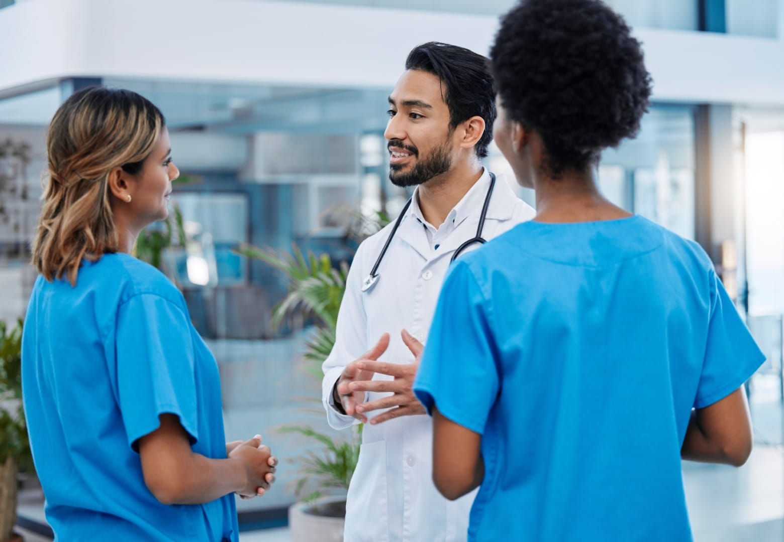 Doctor in a white coat discussing with two nurses in blue scrubs in a hospital setting.