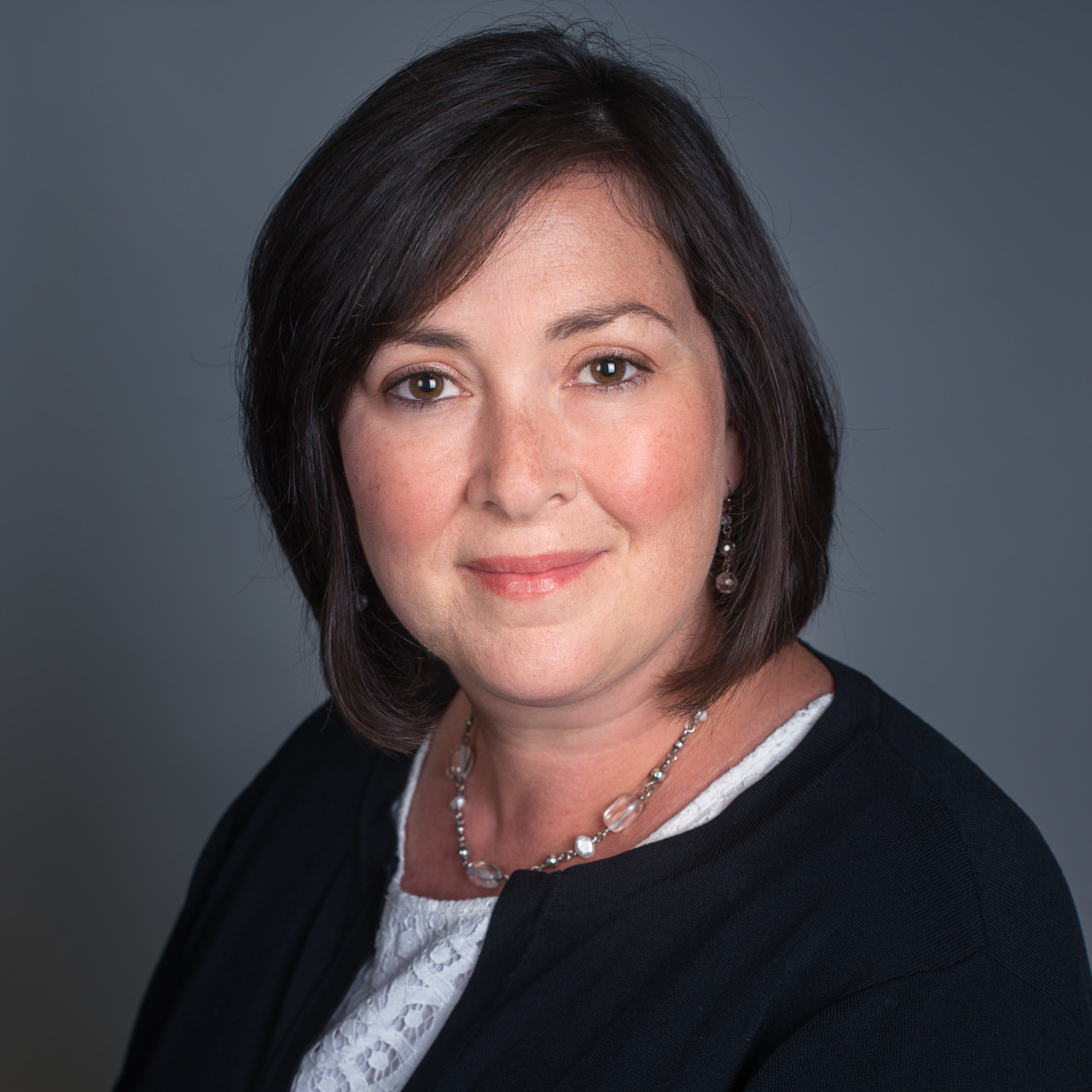 Person with short dark hair wearing a black jacket and necklace, against a gray background.
