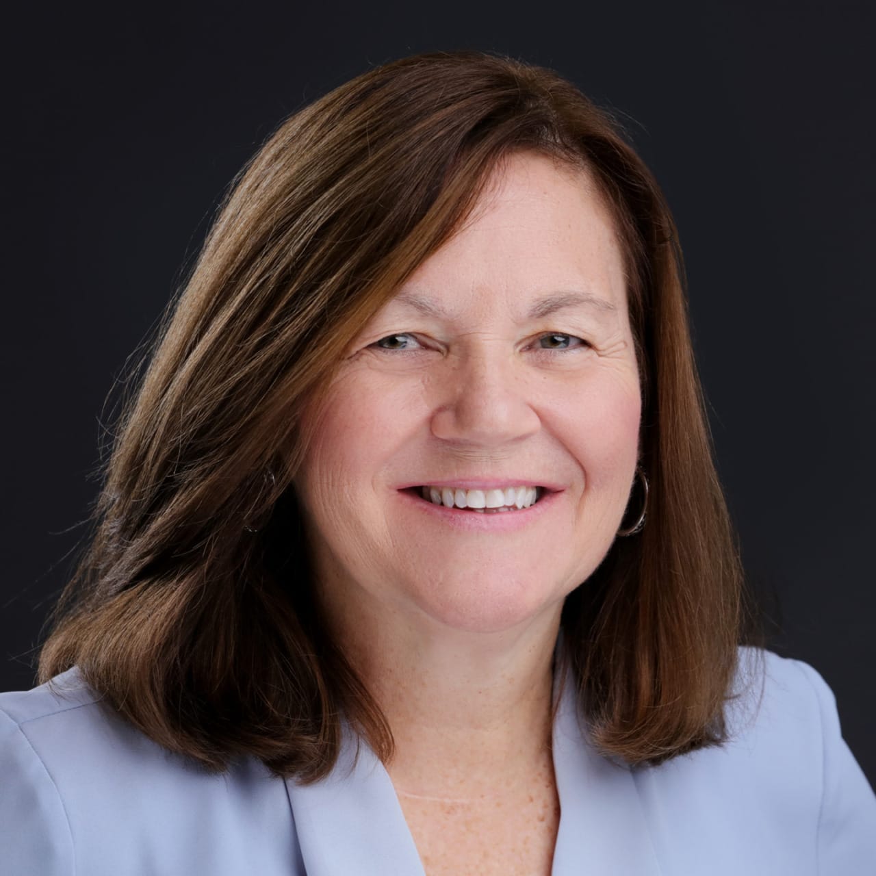 Smiling woman with brown hair wearing a light blue blazer against a dark background.