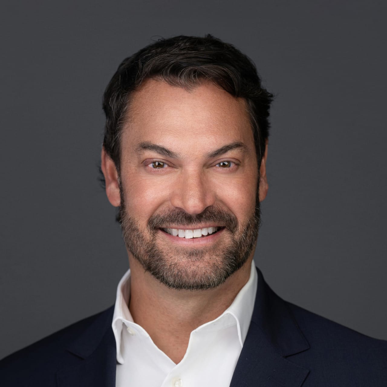 A person with dark hair and beard smiling, wearing a white shirt and dark suit against a plain background.