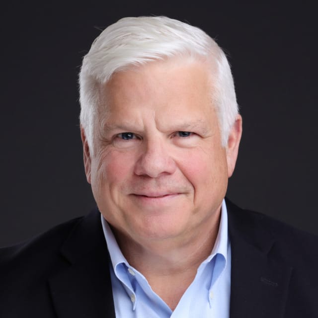 Smiling individual with white hair in a suit jacket and blue shirt against a dark background.