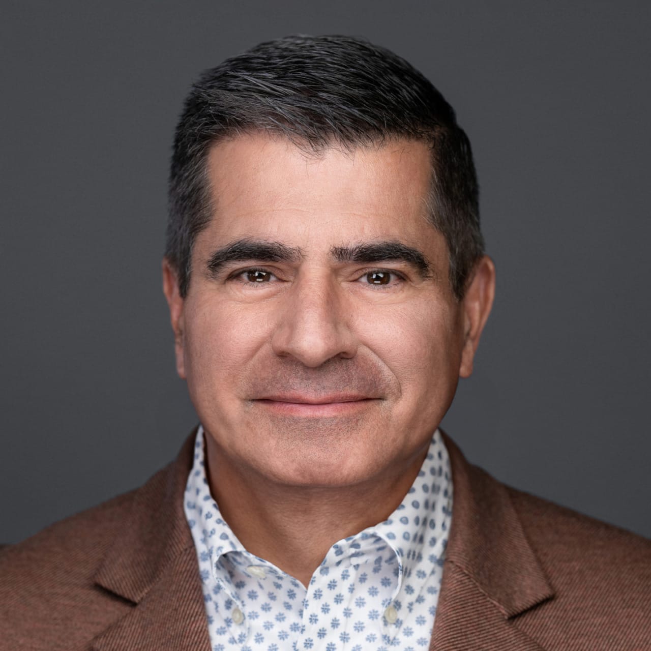 A person in a brown blazer with a patterned shirt, smiling gently against a gray background.
