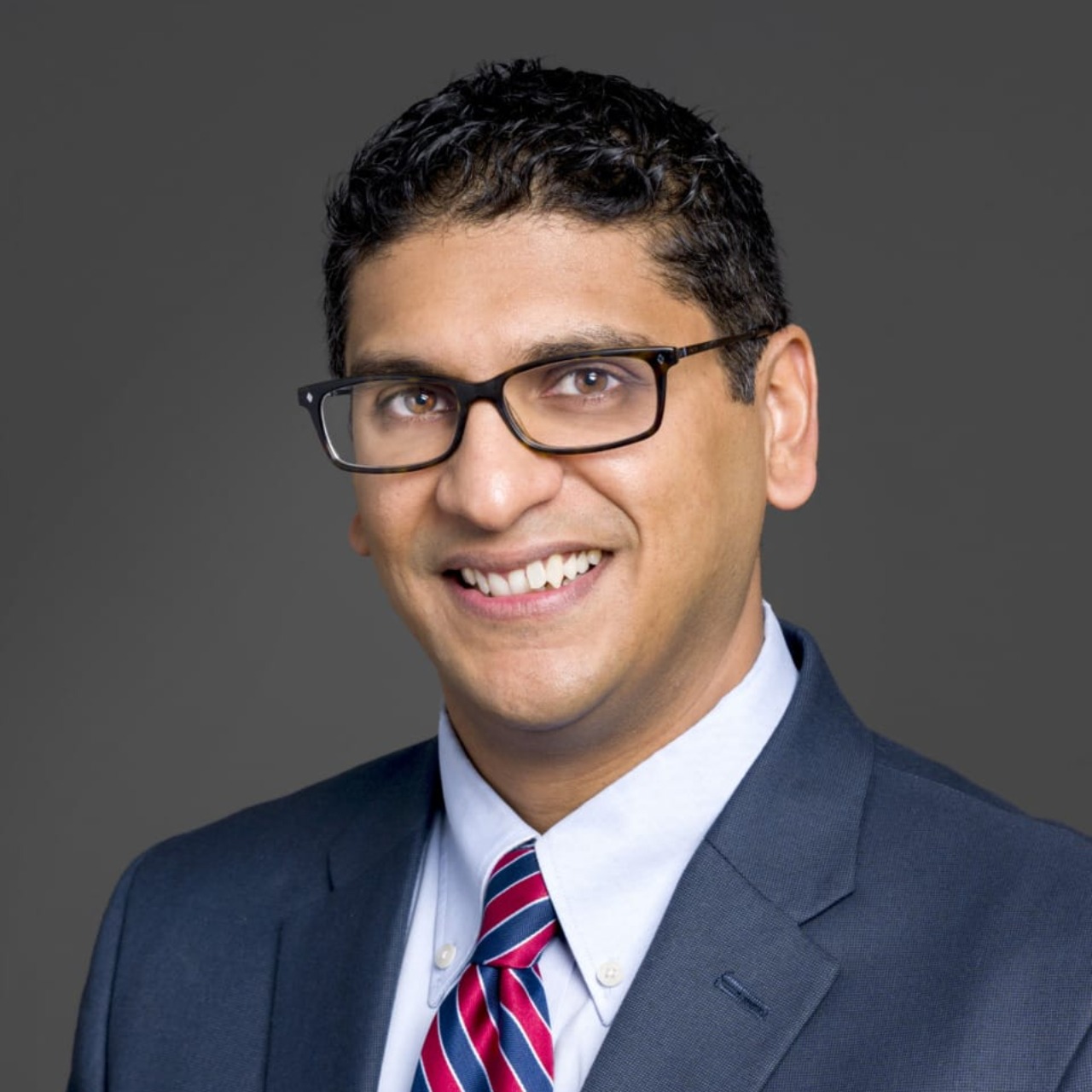 Smiling person in a suit with a red and blue striped tie against a gray background.