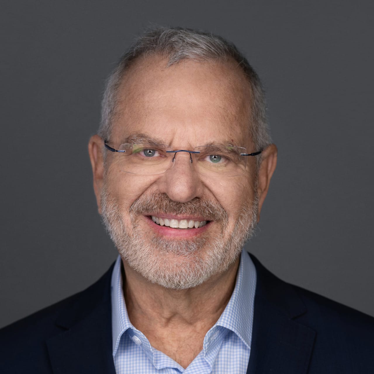Smiling man with gray beard and glasses wearing a suit against a gray background.