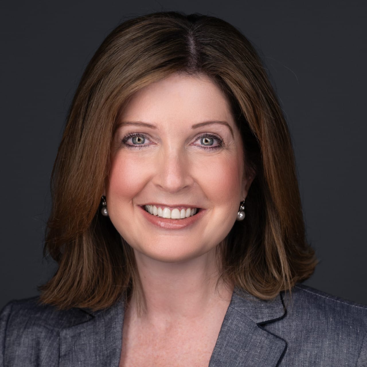 Smiling woman with shoulder-length brown hair wearing a gray blazer against a dark background.