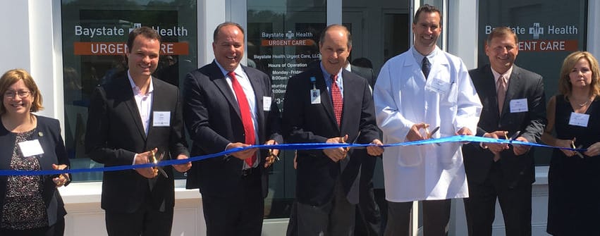 Group of people in suits and a chef's jacket cutting a blue ribbon in front of a building entrance.