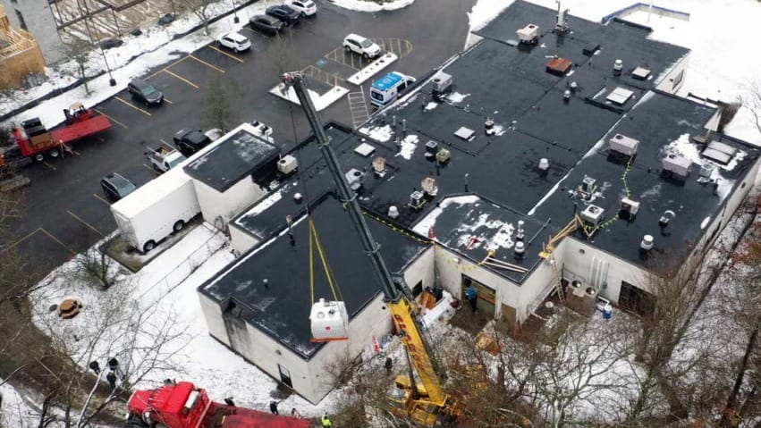 Aerial view of a crane lifting equipment onto the roof of a large building with surrounding parking area in winter.