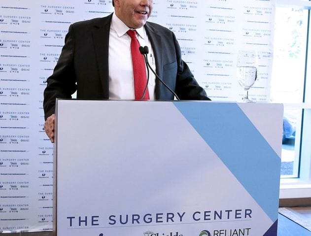 Man speaking at a podium with a sign for The Surgery Center and sponsor logos in the background.