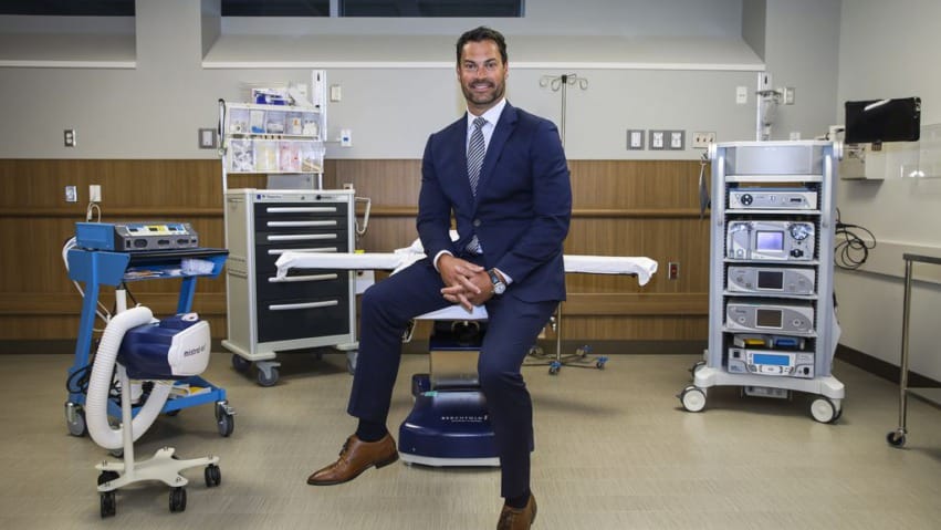 A person in a suit sitting on a stool in a medical office with equipment in the background