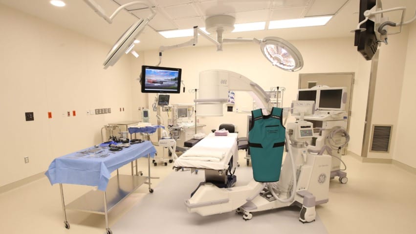 Modern hospital operating room with advanced medical equipment, surgical bed, monitors, and overhead lighting.