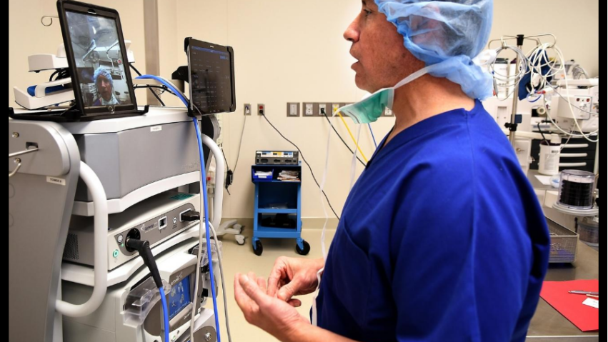 Surgeon in blue scrubs and cap participates in a video call next to medical equipment in an operating room.