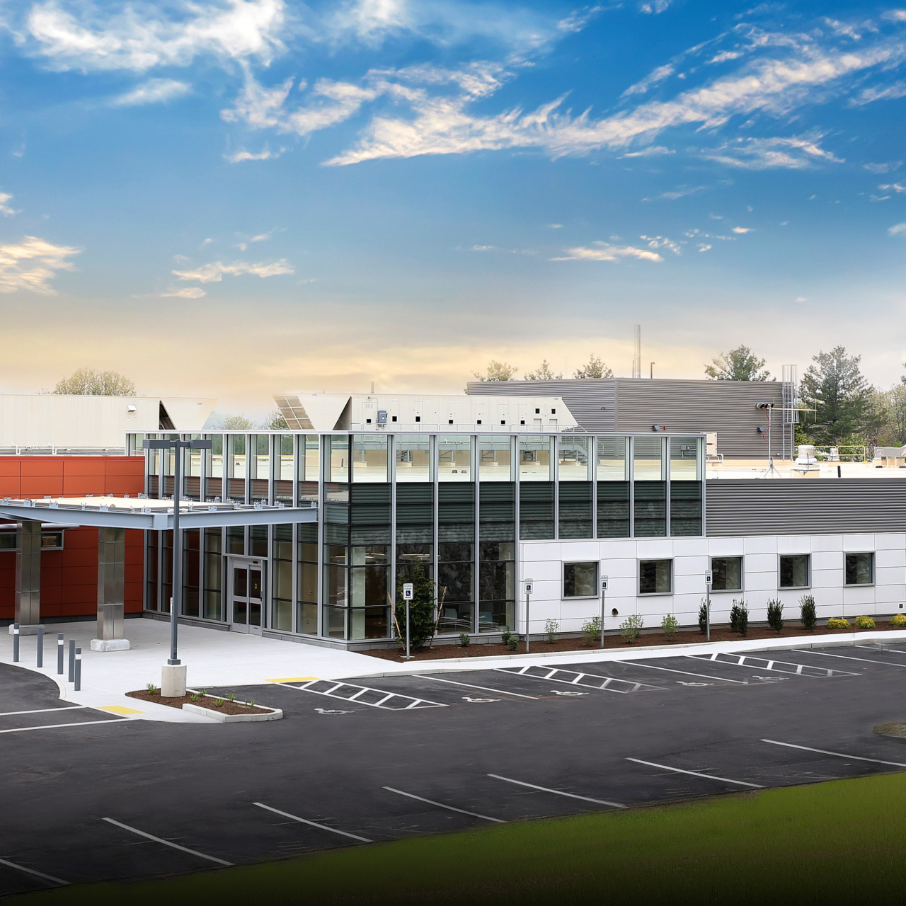 Modern building with glass facade, orange accent, and large parking area, set against a backdrop of trees and blue sky.