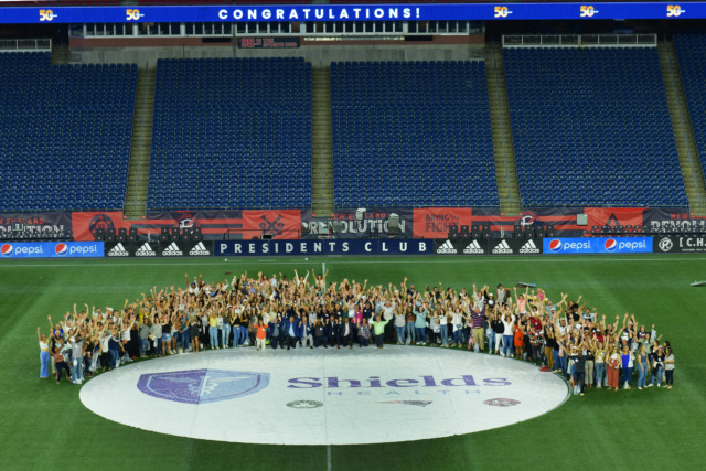 Large group of people gathered on a soccer field, celebrating with a banner overhead that reads "Congratulations!"