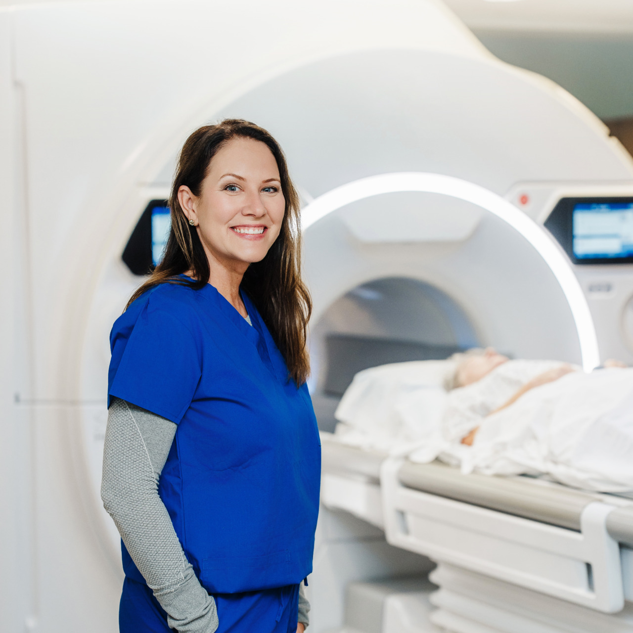 Radiology technician in blue scrubs smiling in front of an MRI machine with a patient undergoing a scan.