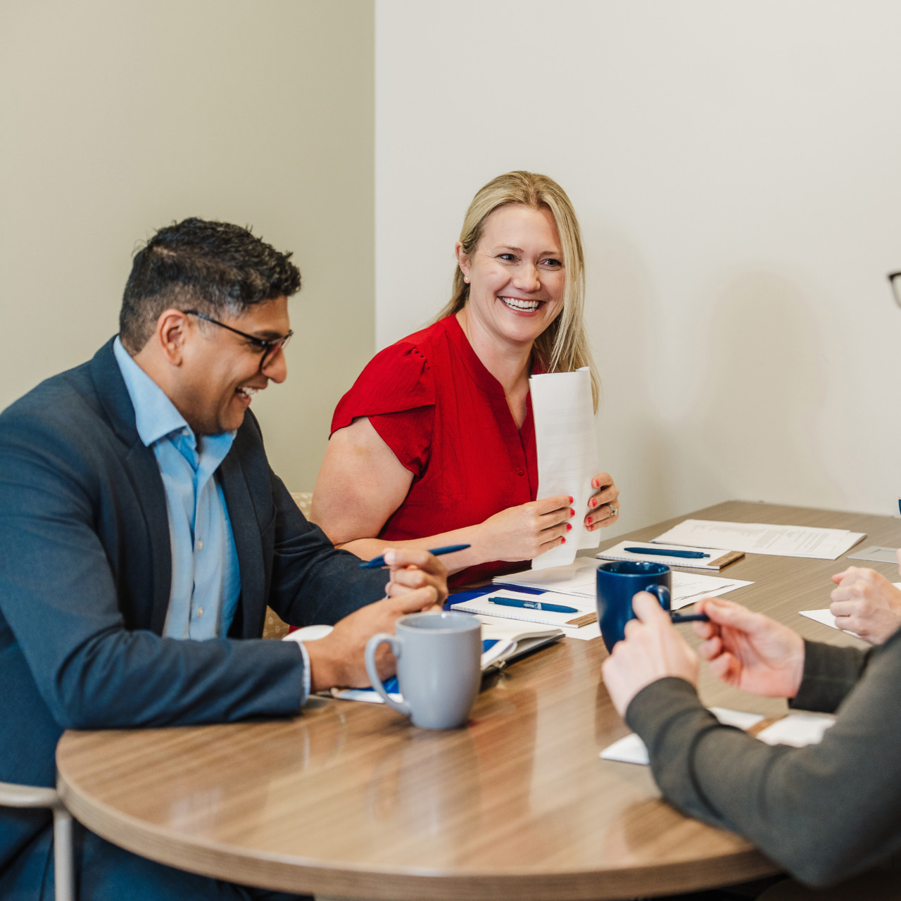 People in a meeting around a conference table, discussing and smiling with papers and coffee mugs in front of them.
