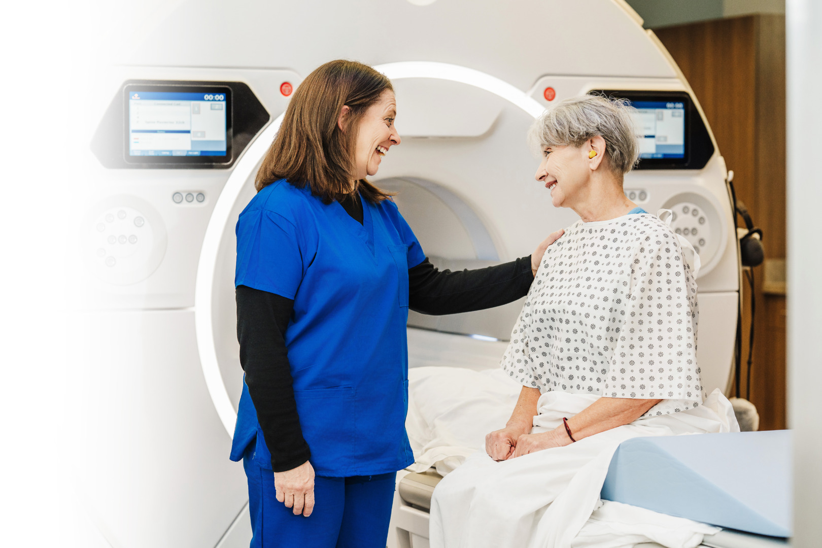 Healthcare professional comforting a patient seated in front of an MRI machine, both smiling and in a clinical setting.