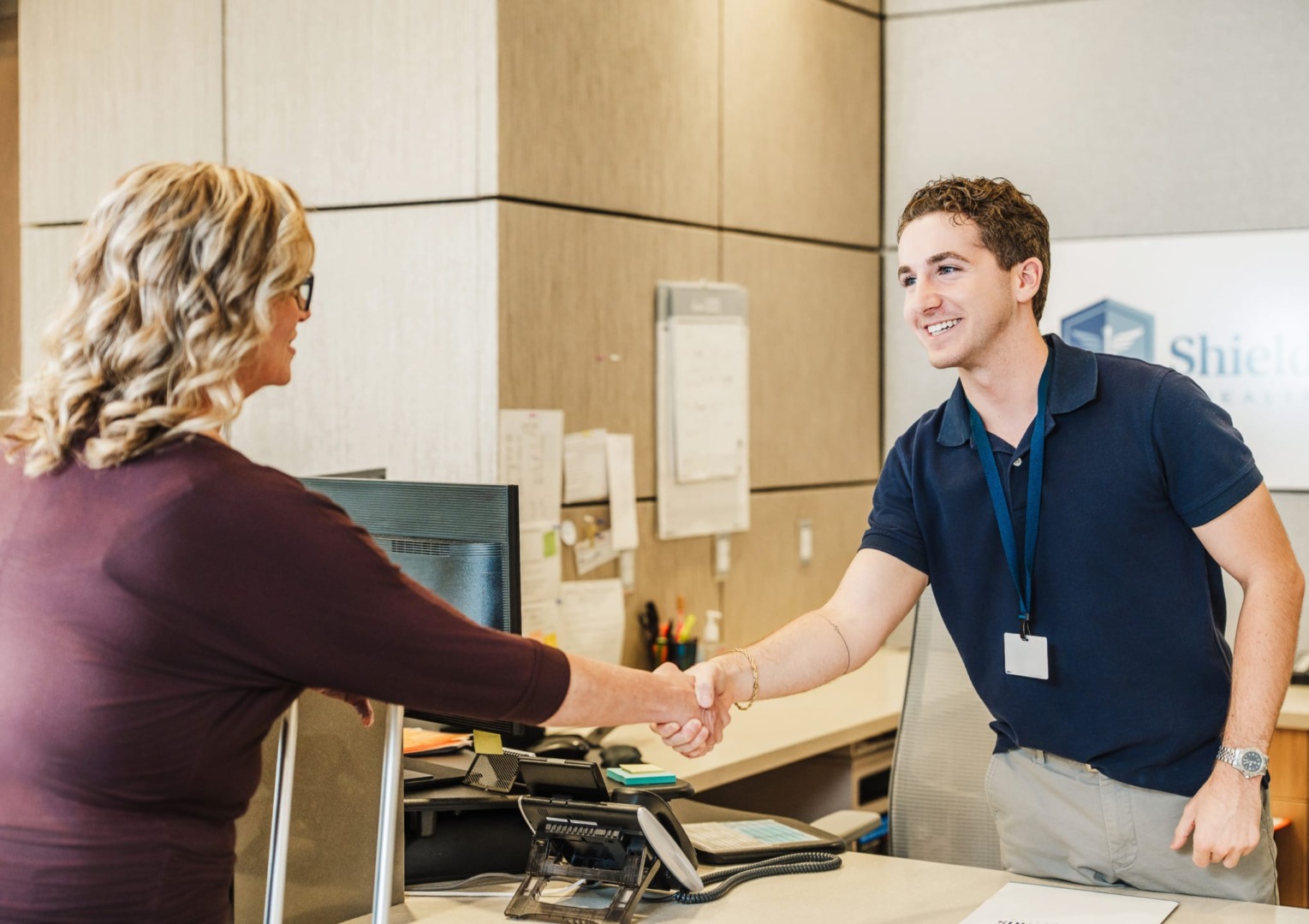Receptionist in a navy blue shirt shakes hands with a woman over a desk in an office setting.