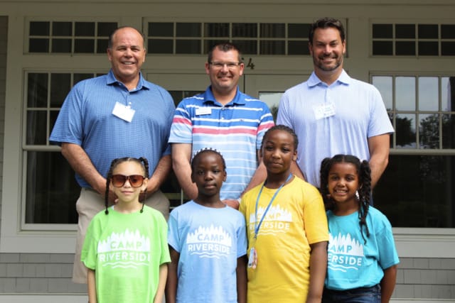 Group photo of three men and four children at Camp Riverside, children wearing colorful camp shirts, standing in front of a building.