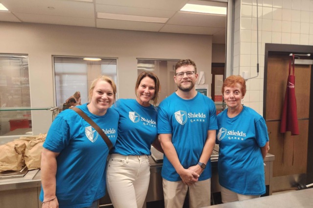 Four people wearing blue Shields Cares shirts standing together in a kitchen setting.