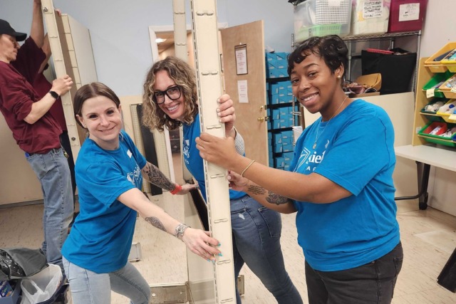 Three people in blue shirts assembling metal shelving in a room, with tools and supplies nearby.