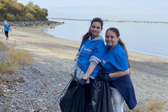 Two people wearing blue shirts clean a beach, holding trash bags and a grabber, with a scenic view of the shoreline.