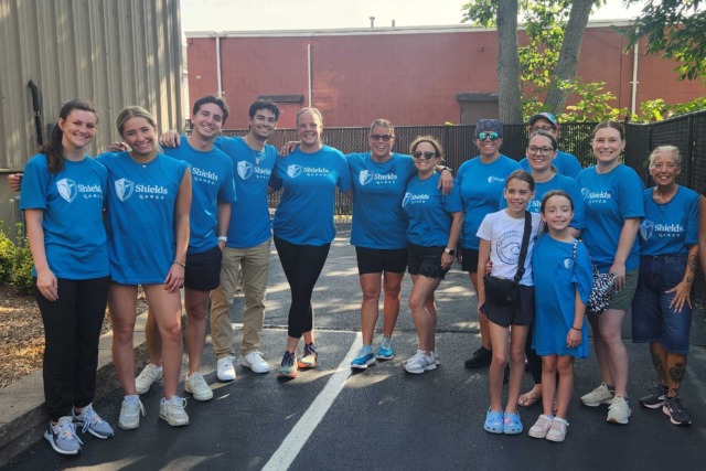 Group of people smiling in matching blue shirts, standing outdoors on a sunny day, posing for a group photo.