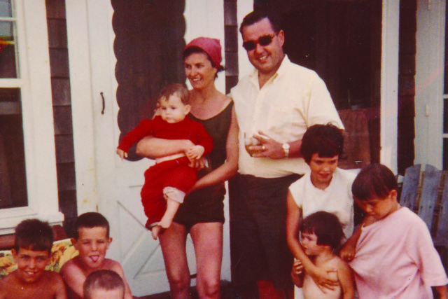 Family posing for a photo outdoors, with children playing and adults standing and smiling.