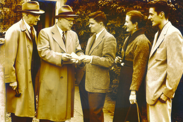 Group of five people in vintage attire engaged in conversation outdoors, holding papers and looking at each other.