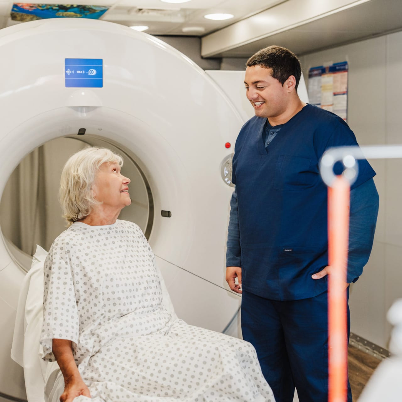 Elderly woman in hospital gown talking with medical professional in blue scrubs near medical imaging machine.