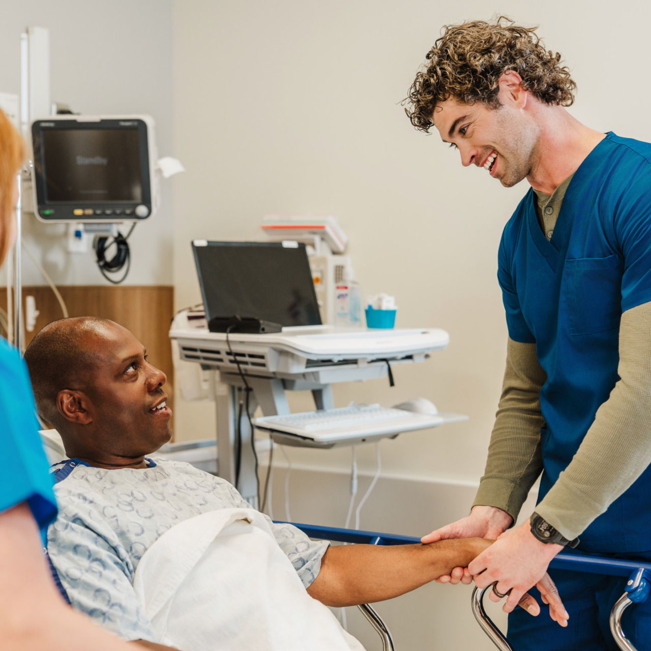 Nurse smiling and talking with a patient in a hospital room, with medical equipment in the background.