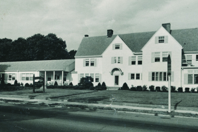 Black and white photo of a large, two-story house with a sloped roof and surrounding trees.