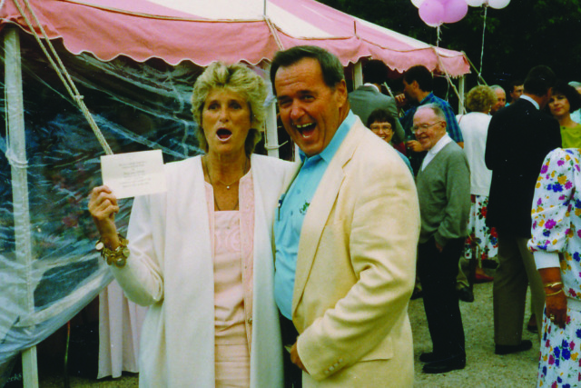 A cheerful couple poses in front of a pink and white tent at an outdoor event with other people in the background.