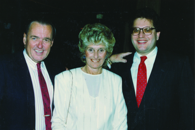 Three people in formal attire smiling at an event with dark background.