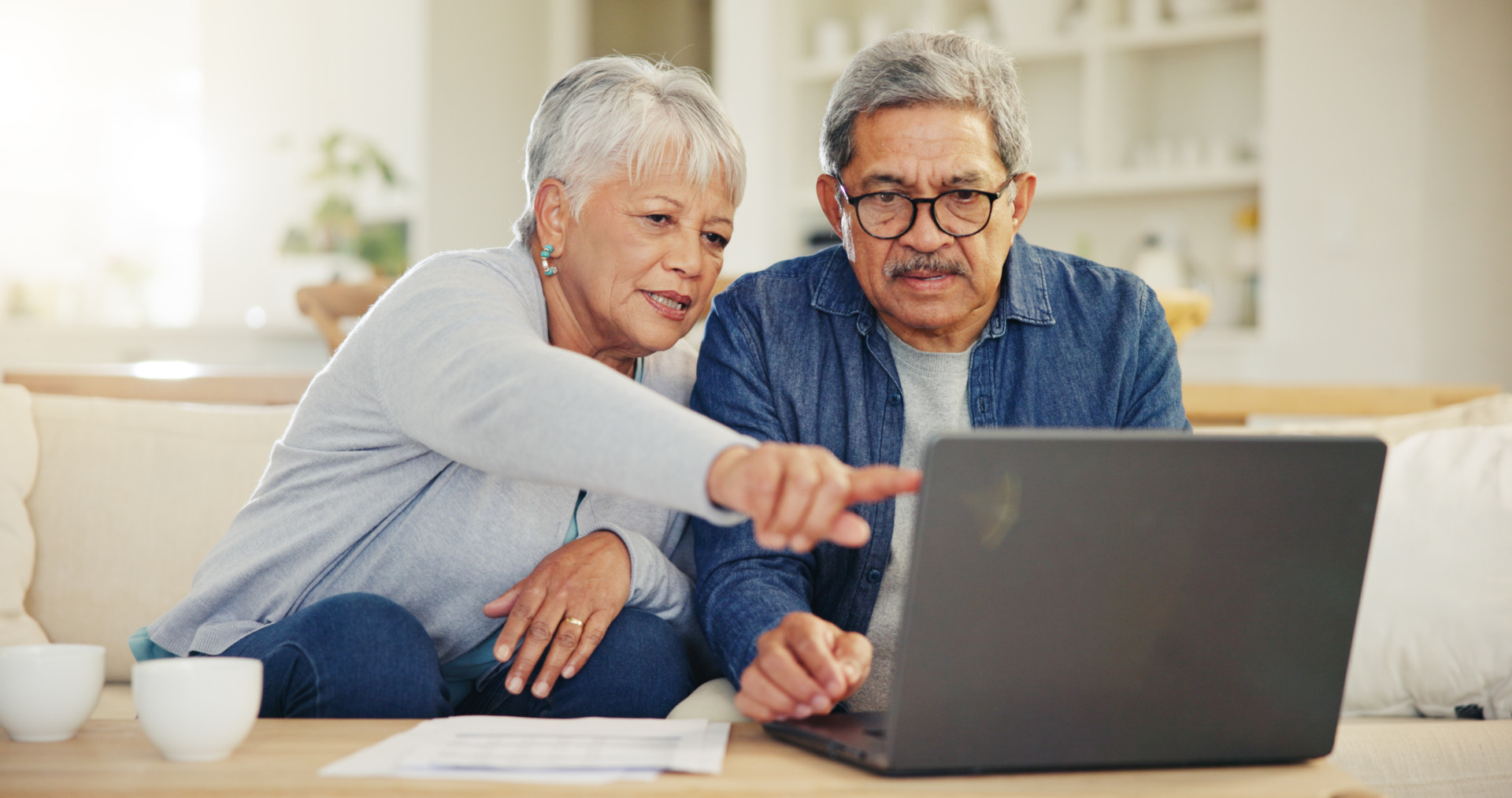 Elderly couple sitting on a couch, focused on a laptop screen, while the woman points and talks, cups on the table nearby.