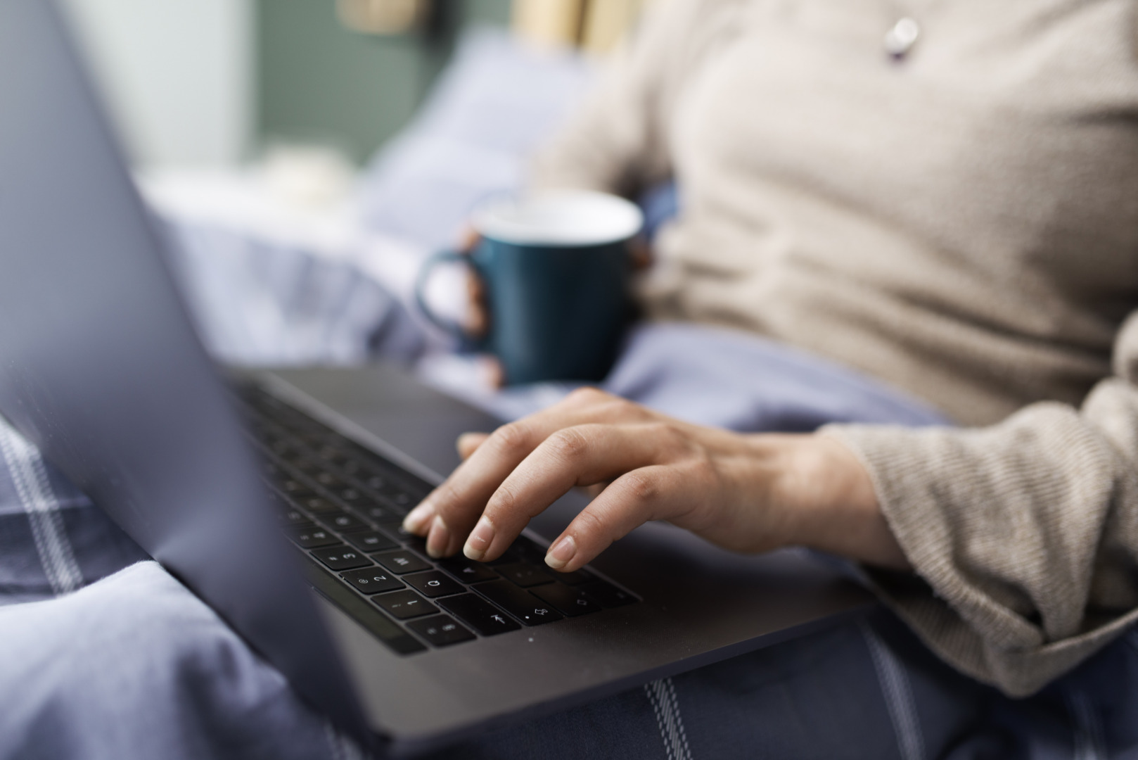 Person using a laptop while holding a green mug, sitting on a bed with blue bedsheet.