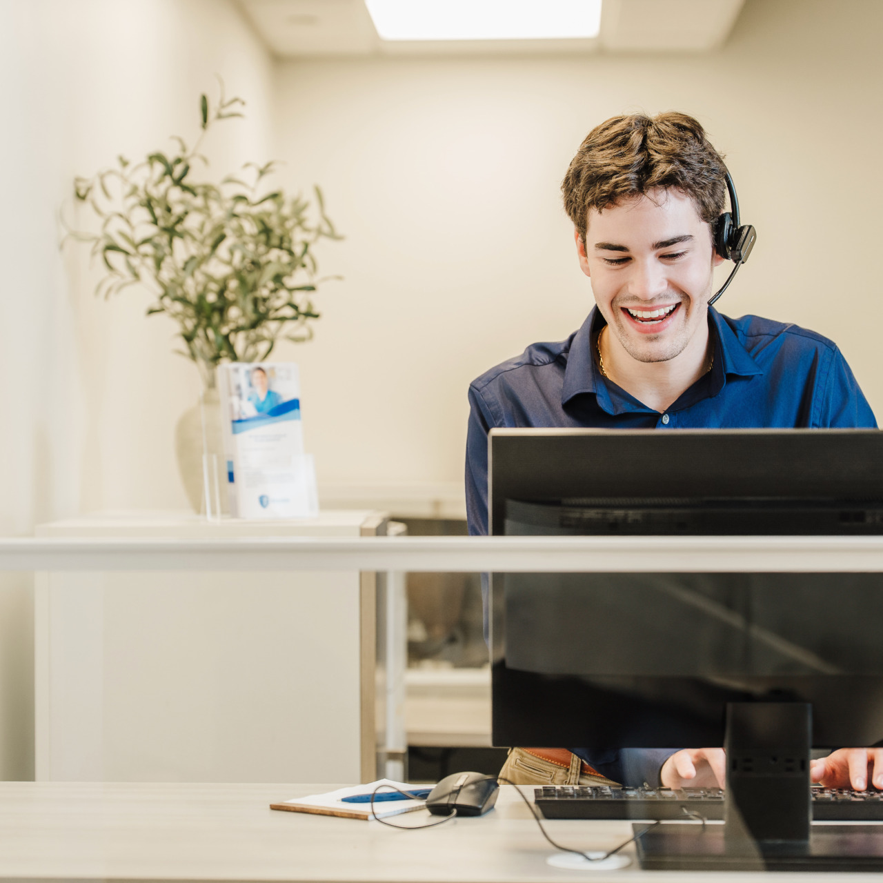 Smiling customer service representative with a headset working at a computer desk in a bright office setting.