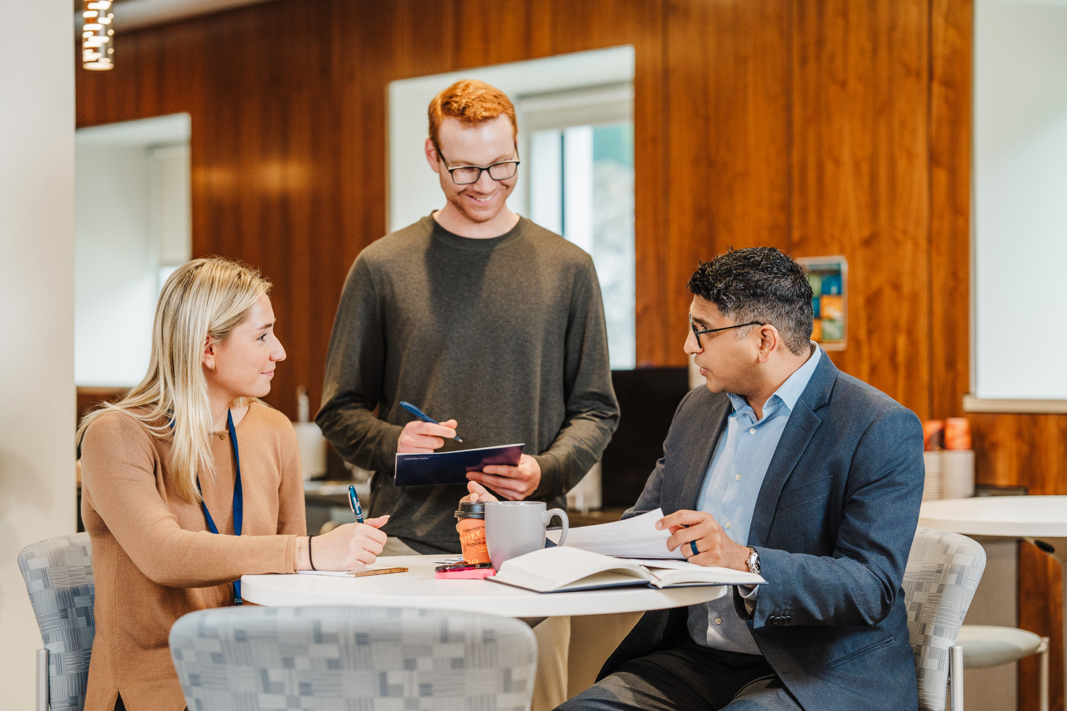 Three people discussing around a table in a modern office setting with notebooks and coffee cups.