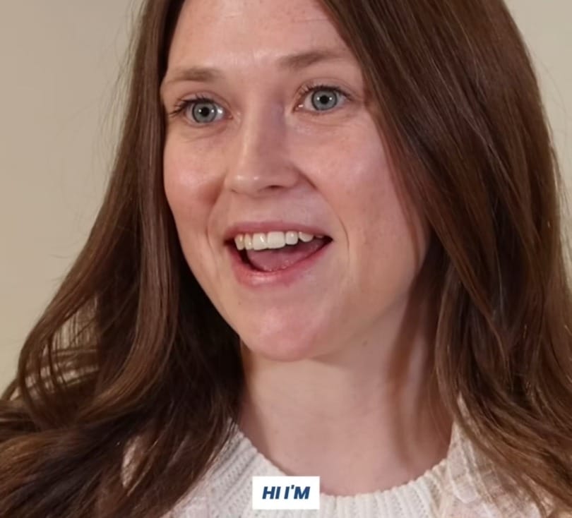 Smiling woman with long brown hair wearing a white sweater and beige scarf in a neutral beige background setting