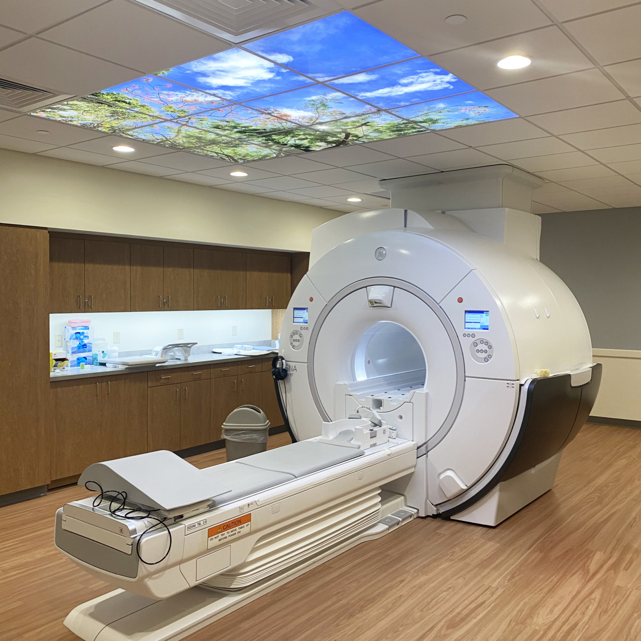 MRI machine in a modern medical room with wood flooring and a sky-themed ceiling panel.