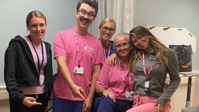 Group of five people wearing pink-themed outfits in a room, smiling and showing off a pair of socks with patterns.
