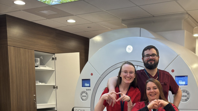 Three medical professionals in red scrubs smiling and making heart shapes with their hands in front of CT machine.
