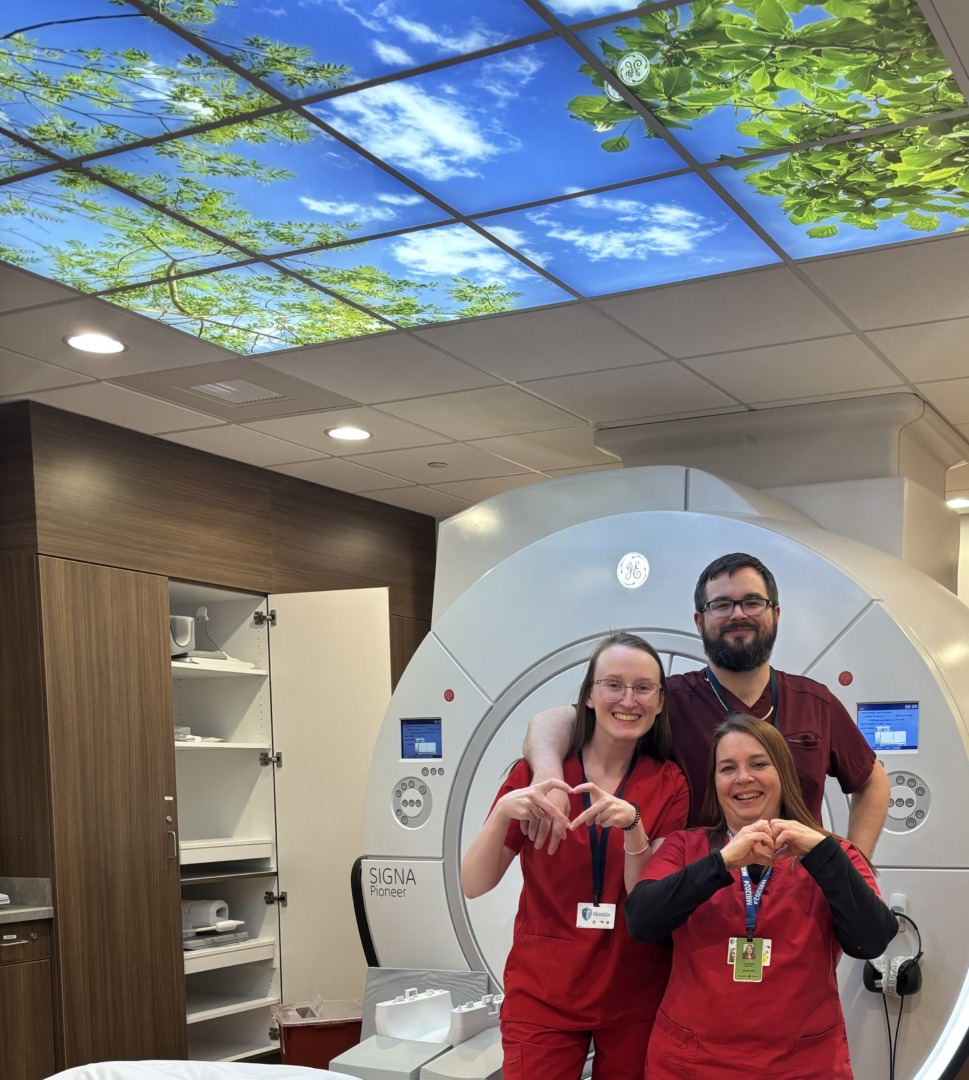 Three medical professionals in red scrubs smiling and making heart shapes with their hands in front of CT machine.